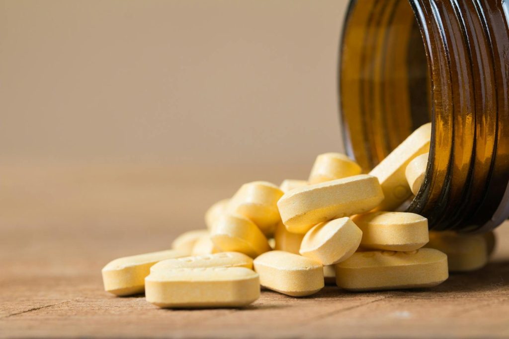 A close-up image of vitamin tablets spilling out of a transparent brown bottle on a wooden surface.