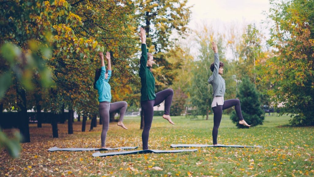Three women practicing yoga outdoors on mats amidst autumn foliage in a park.