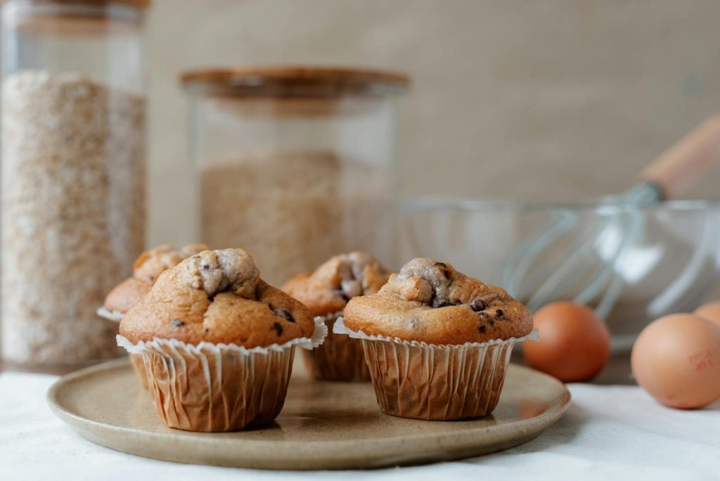 Delicious muffins with soft texture and chocolate chips near containers with ingredients and raw eggs