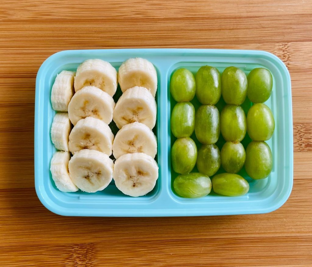 A healthy snack of sliced bananas and green grapes in a blue plastic container on a wooden table.