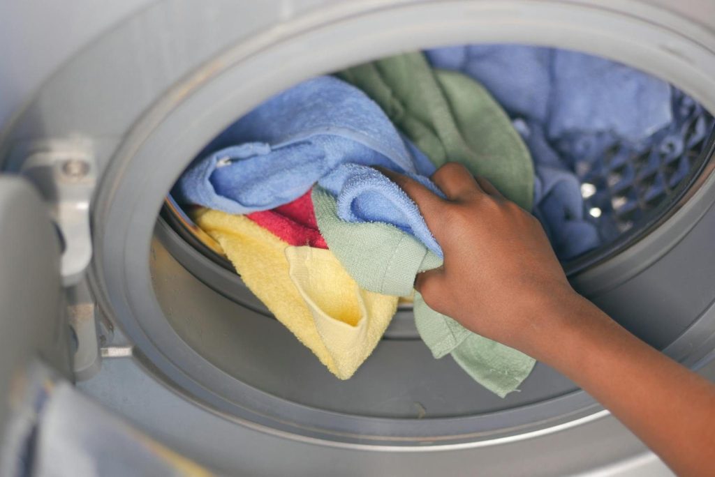 A close-up of a hand loading colorful towels into a front-loading washing machine.
