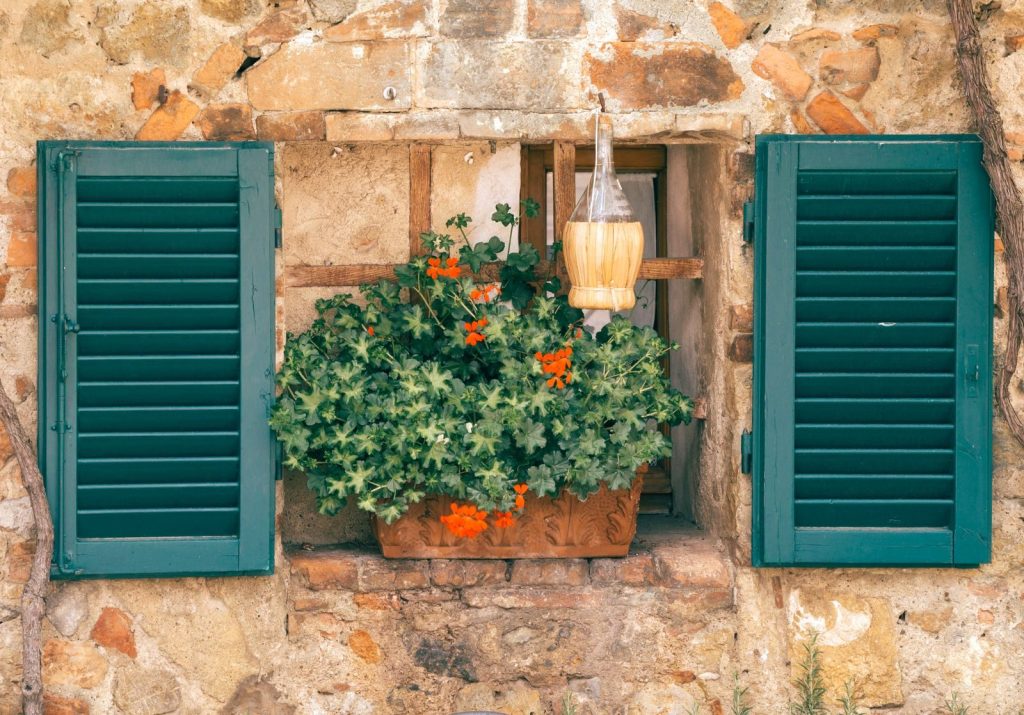 Charming rustic stone wall window with vibrant flowers and green shutters.