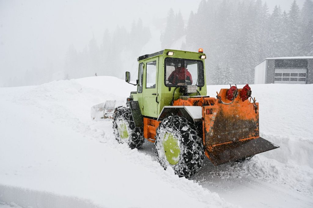 Green and orange snowplow clearing heavy snow in a snowstorm on a frosty day.