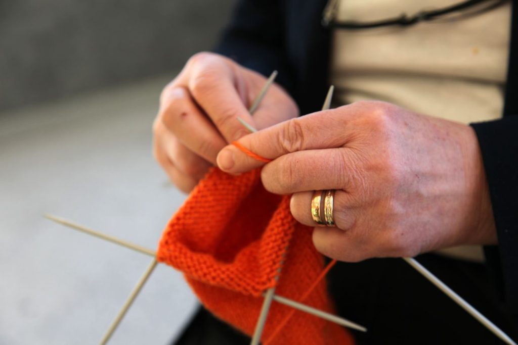A close-up of hands knitting a bright orange fabric with needles, showcasing skillful craftsmanship.