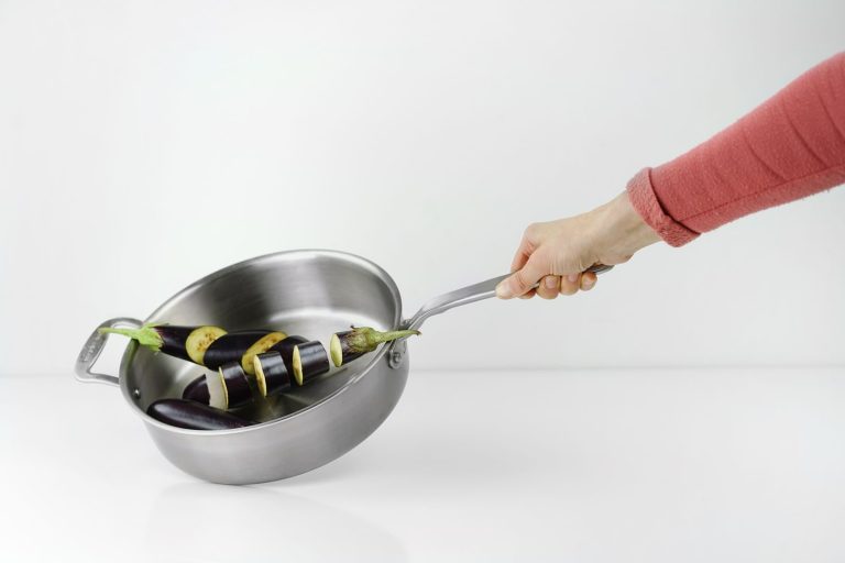 A hand holding a skillet with freshly sliced eggplant, against a white background.