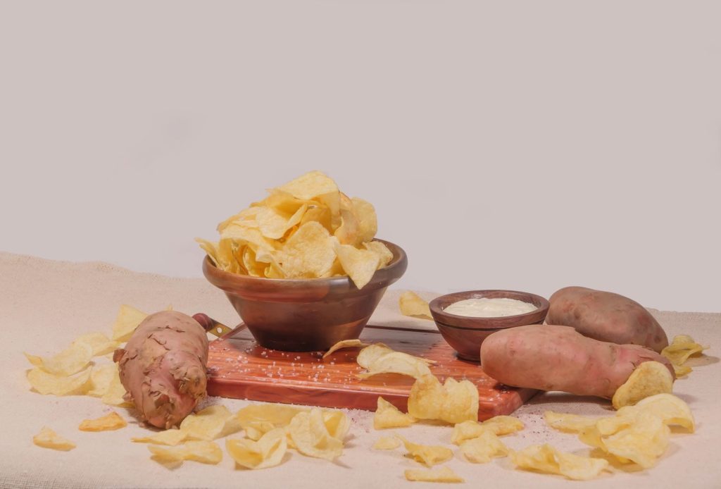 A wooden bowl of crispy potato chips with a creamy dip and whole potatoes on a rustic tray.
