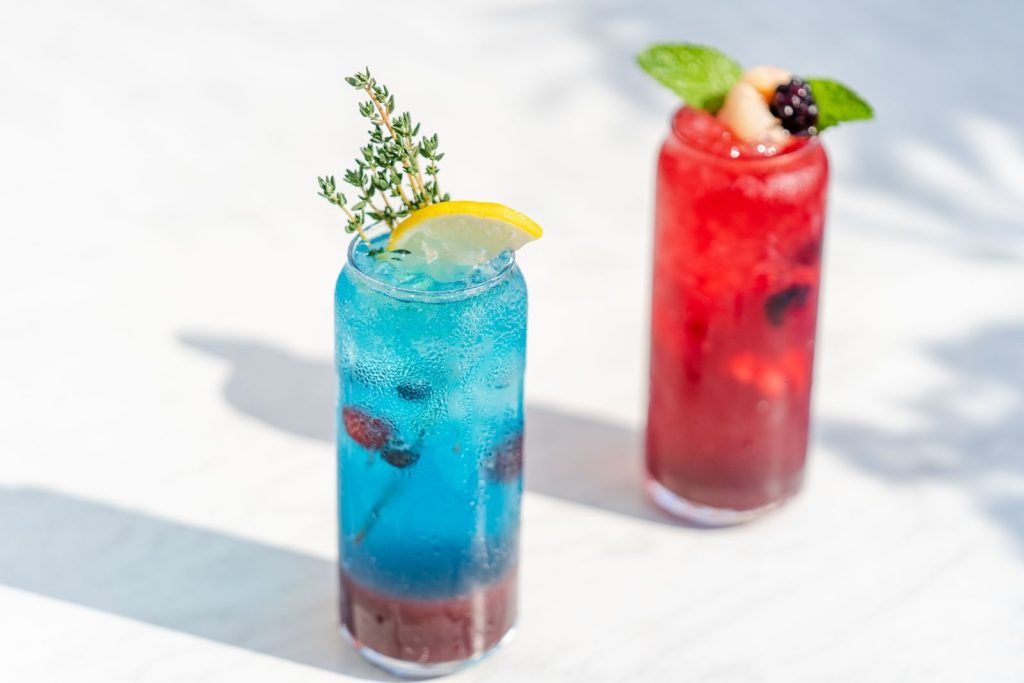 Vibrant blue and red cocktails garnished with fresh herbs and berries on a sunlit table.