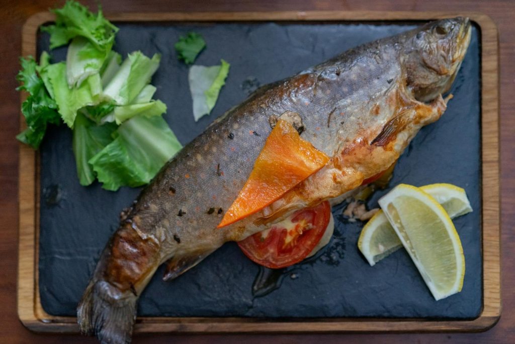 Close-up of a whole grilled fish served with lemon slices and greens on a slate tabletop.