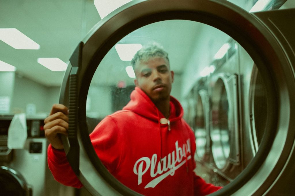 A young man in a red hoodie is reflected through a washer door at a laundromat.