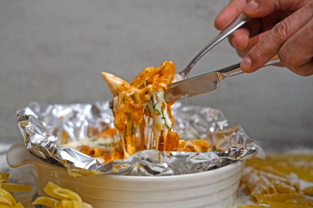 Delicious cheesy baked pasta served in foil with fresh herbs and a hand holding a fork and knife.