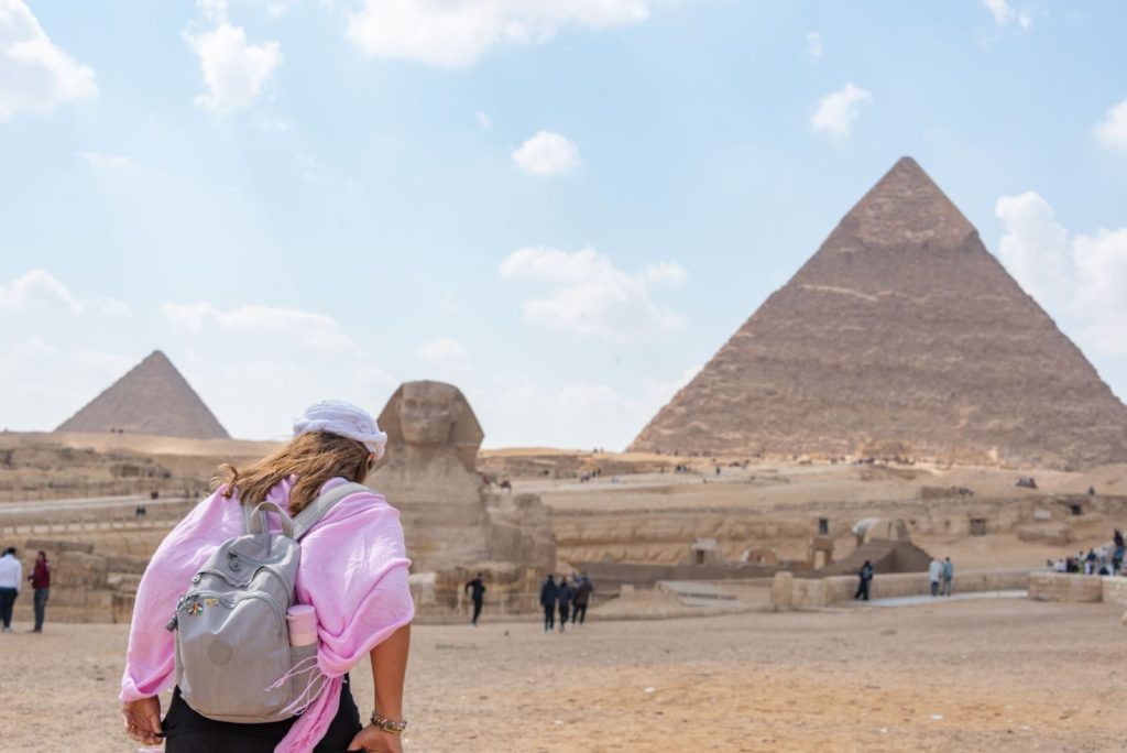 A tourist in a pink shirt wanders the Pyramids of Giza and Sphinx under a clear sky.
