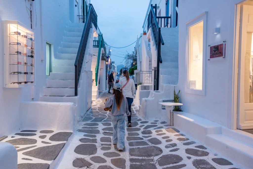 People walking through a picturesque Greek alley with traditional white architecture at dusk.