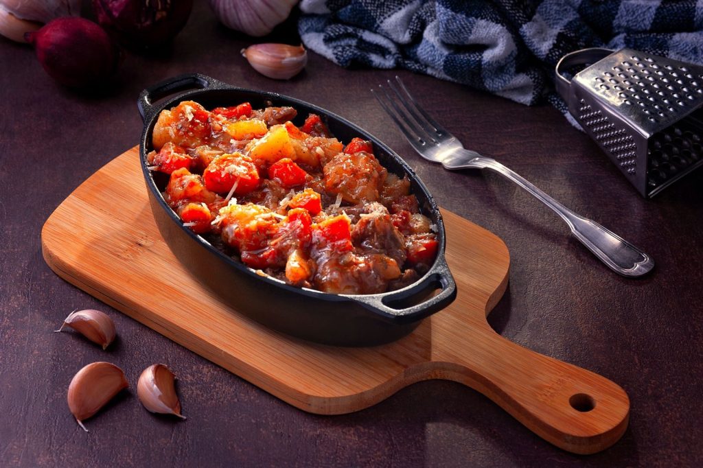 Savory vegetable stew served in cast iron on wooden board, paired with garlic cloves and kitchen utensils.