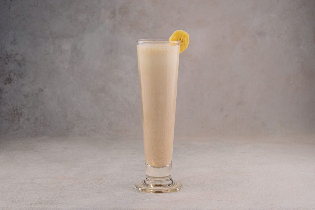 Tall glass of banana smoothie with a banana slice garnish against a neutral textured background.