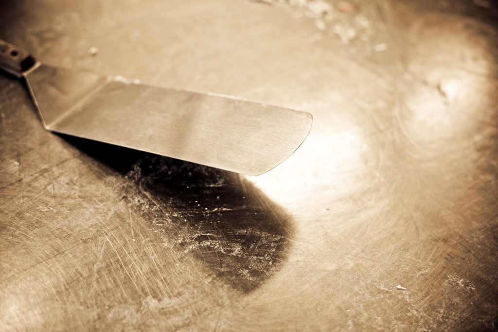 Close-up of a vintage stainless steel spatula on a worn countertop with sepia tones.