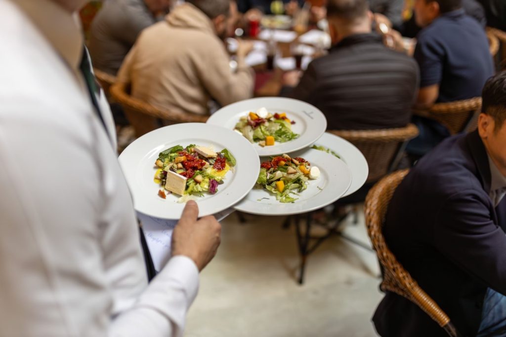 Waiter carrying plates with fresh salads in a busy restaurant setting with diners in background.