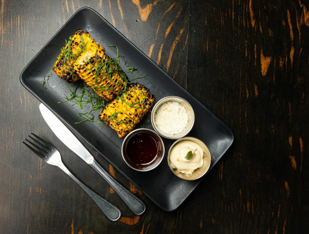 Appetizing grilled corn with a variety of dipping sauces served on a dark wooden table.