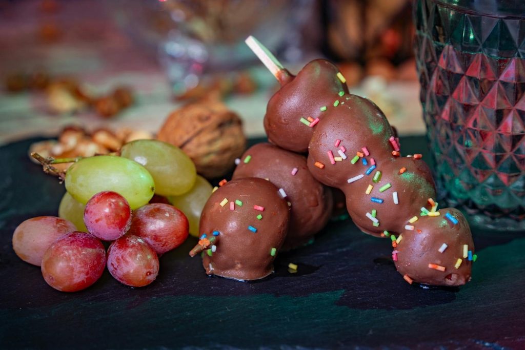 Still life showcasing chocolate-dipped fruit skewers with nuts and grapes on a slate board.