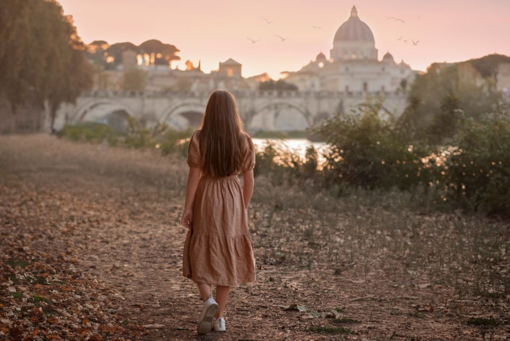 A young woman walks along a path with St. Peter's Basilica in the background during sunset in Rome.