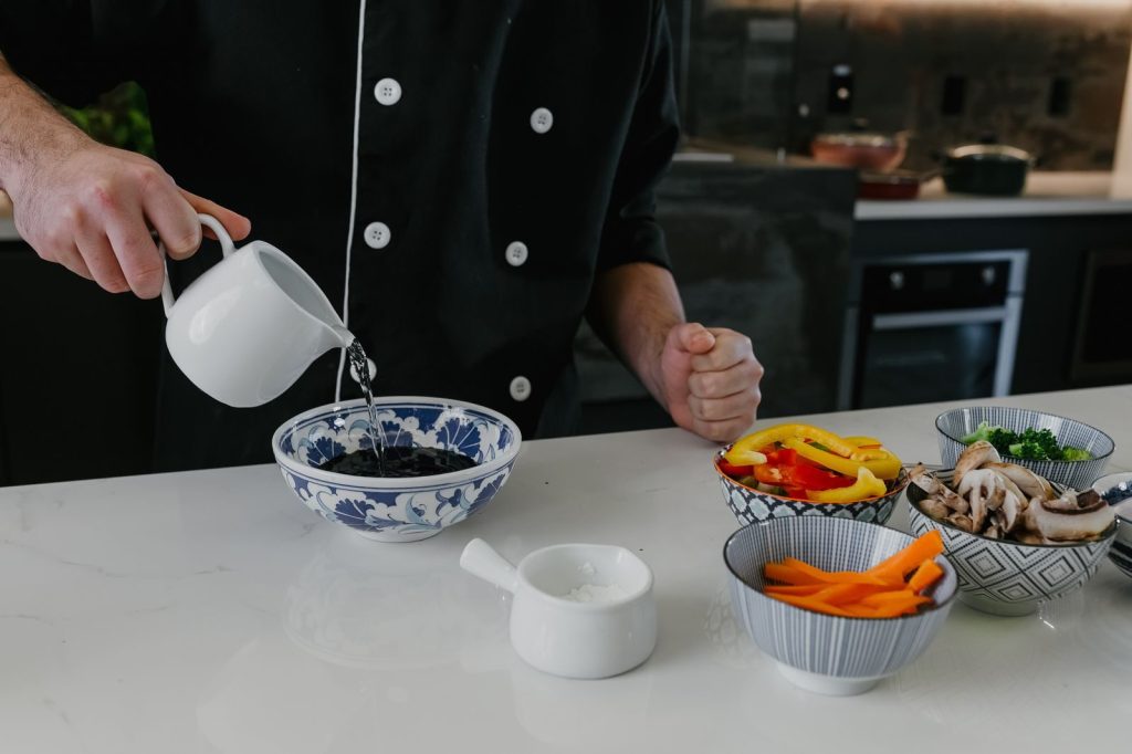 A chef prepares vegetables and pours water into a bowl in a modern kitchen setting.