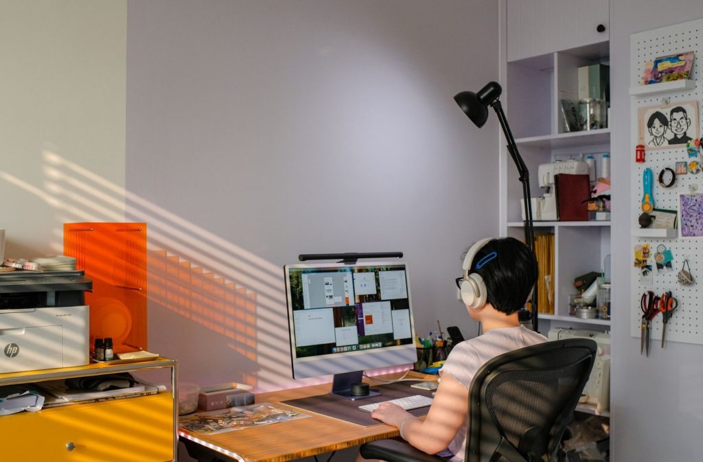 An individual with headphones working on a desktop computer in a home office setting during the day.
