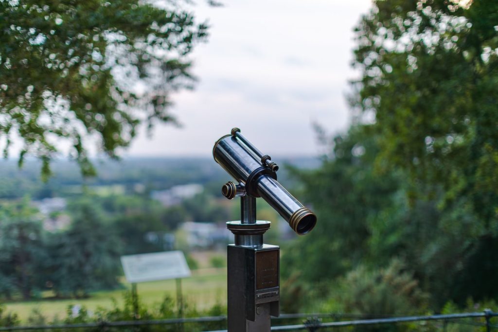 A public telescope pointing towards a scenic green landscape during the day.