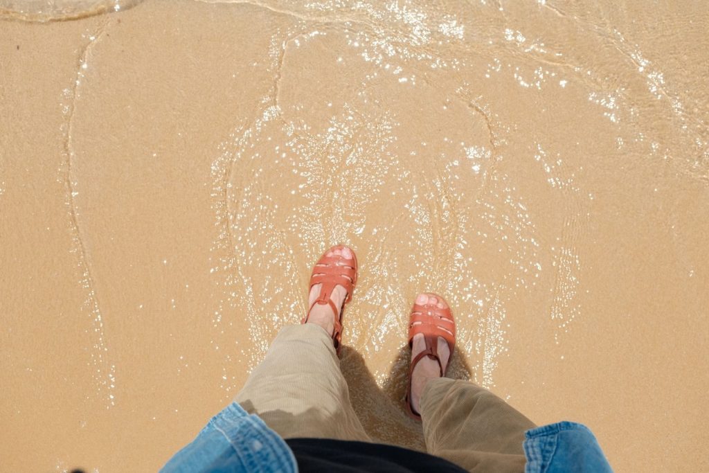 Person standing in sand with water, wearing sandals and casual clothing, on a sunny beach.