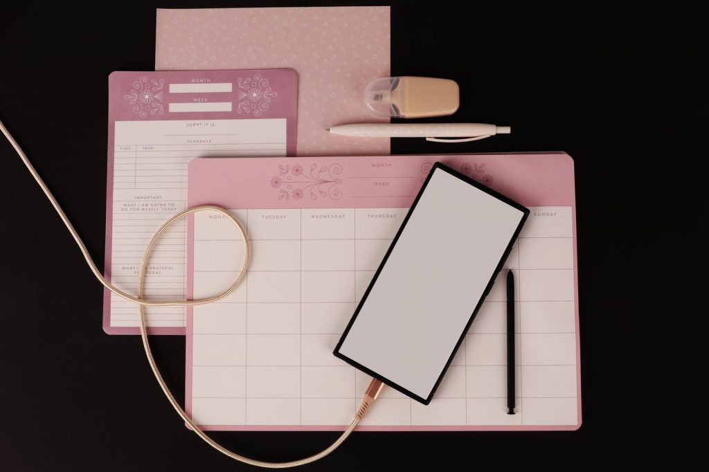 Flat lay of a desk with planner, tablet, pen, and stationery on black background.