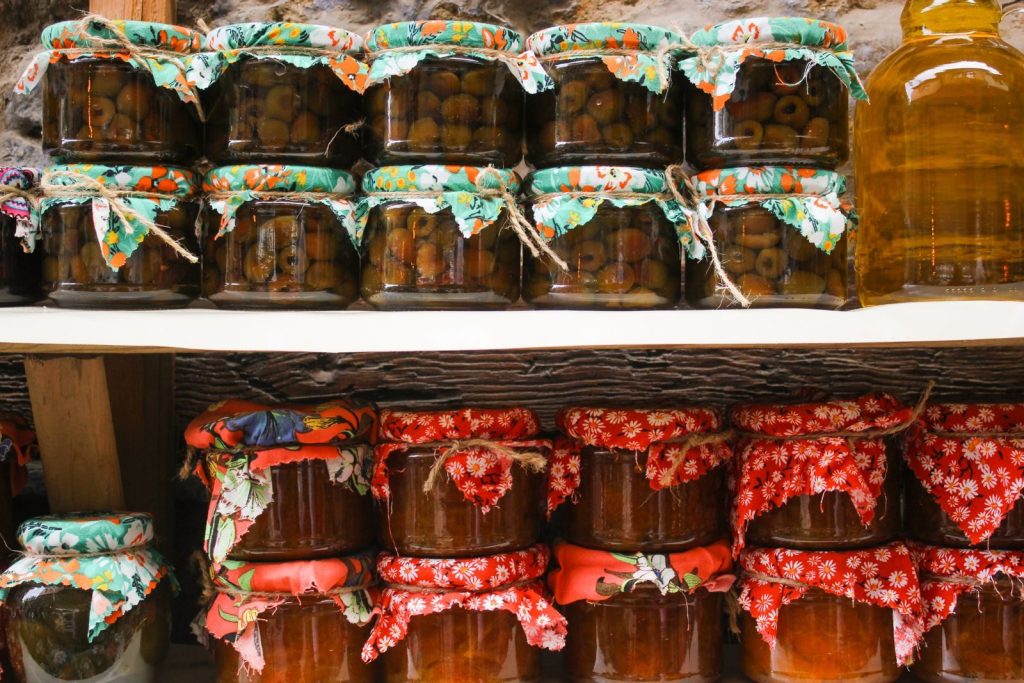 Variety of colorful homemade preserves and pickles in jars with fabric covers.
