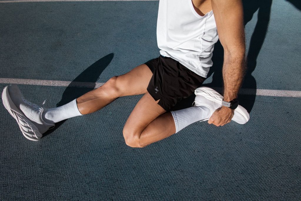 Male athlete stretching on an outdoor track, wearing white sneakers and black shorts for fitness.