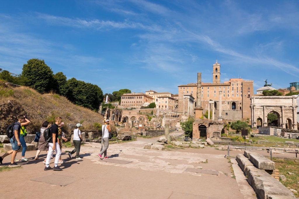 Visitors walking through the historic Roman Forum with ancient ruins and buildings under a clear blue sky.