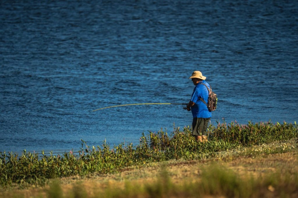 An adult male fishing by a serene lake, wearing a hat and blue shirt.
