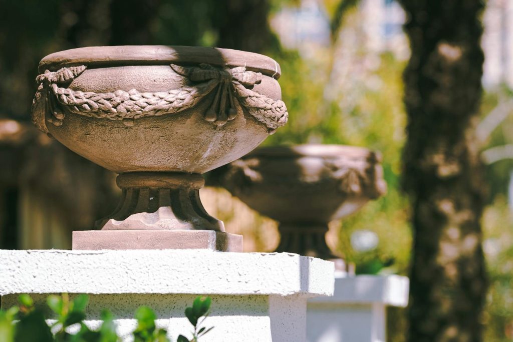 Stone urns with intricate details displayed on white pedestals in a sunny outdoor setting.