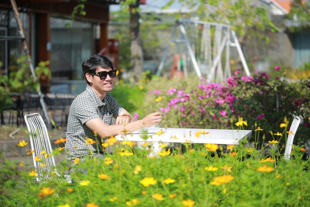 Man relaxing at a garden café with flowers in bloom under a clear sky.