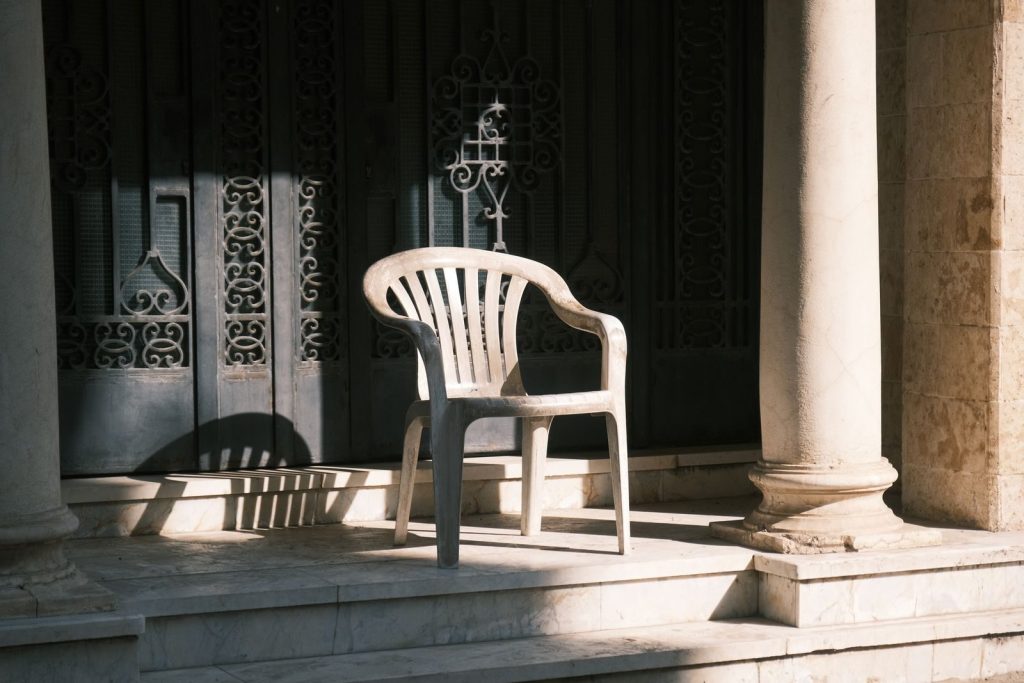 A lone plastic chair sits quietly under sunlight on a patio with ornate doors.