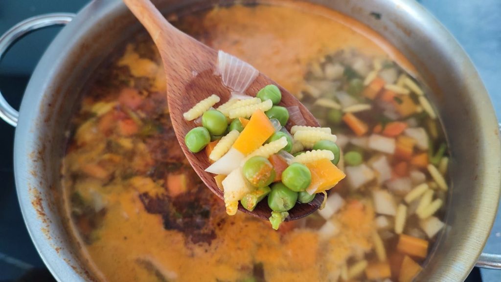 Close-up of a wooden spoon holding fresh vegetables over simmering soup.