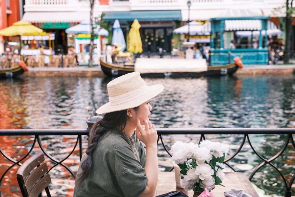 A woman in a hat sits by a riverside cafe, enjoying the view on a summer day.