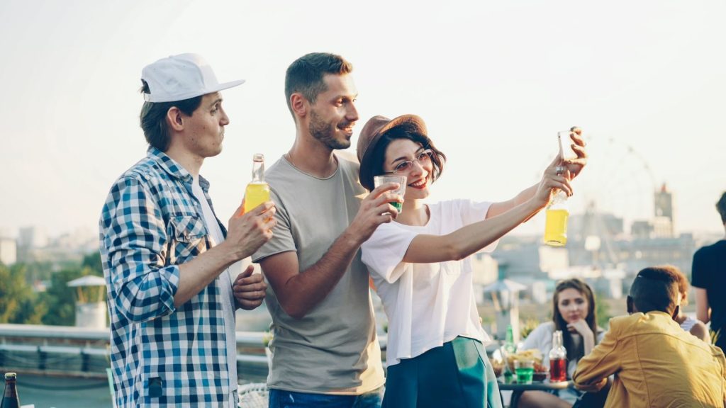 Three friends taking a selfie at a rooftop party with drinks, city view background.