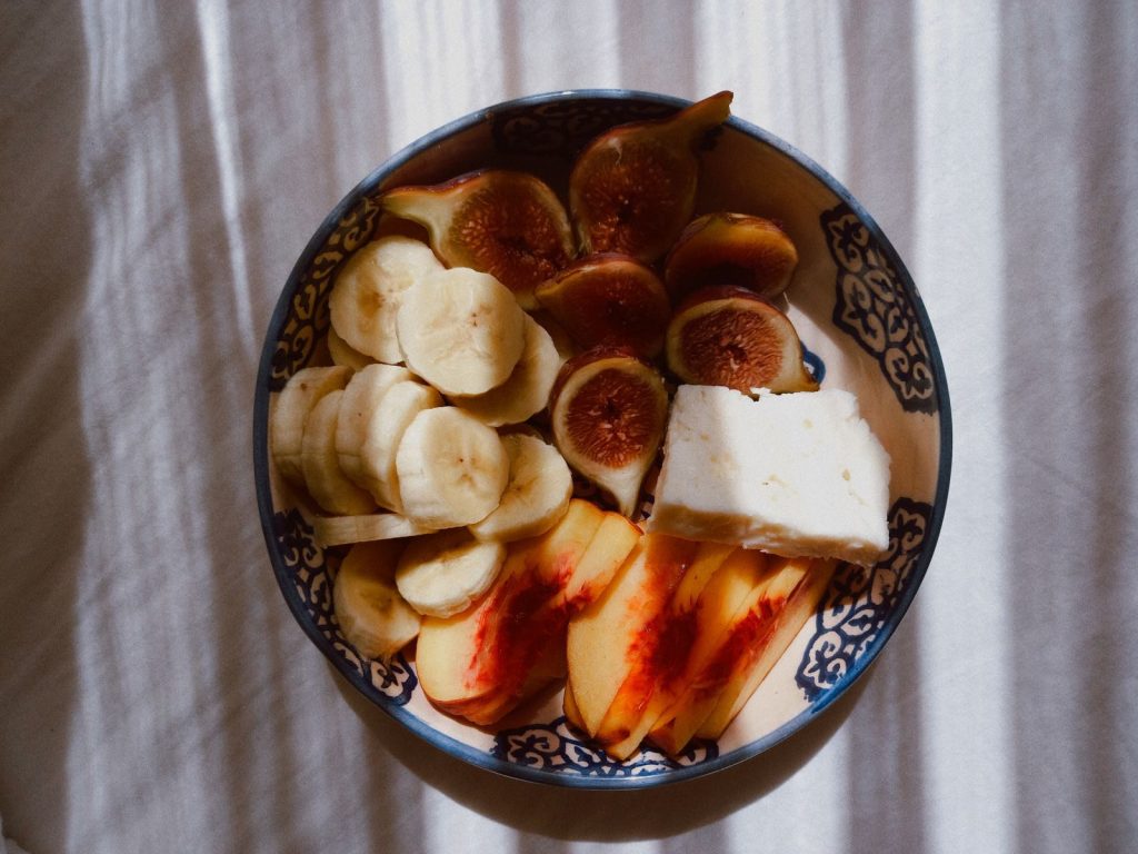 A vibrant bowl of banana, fig, peach, and cheese in decorative dish under sunlight shadows.