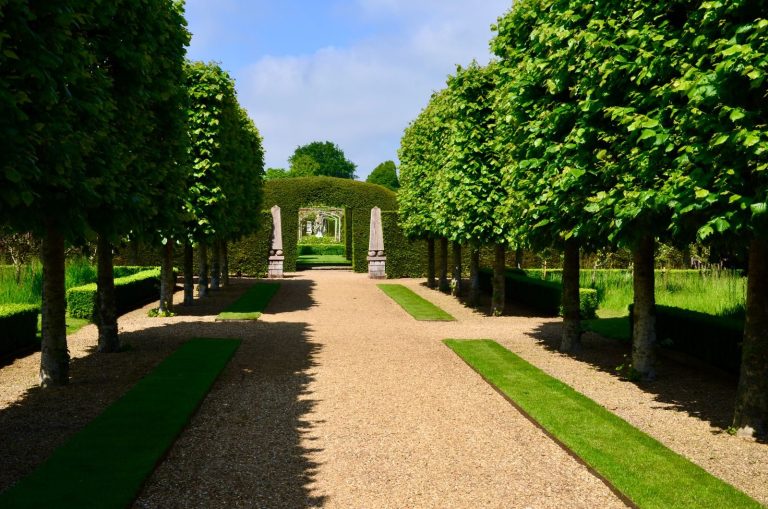 A picturesque garden pathway flanked by symmetrical trees and hedges under a clear sky.