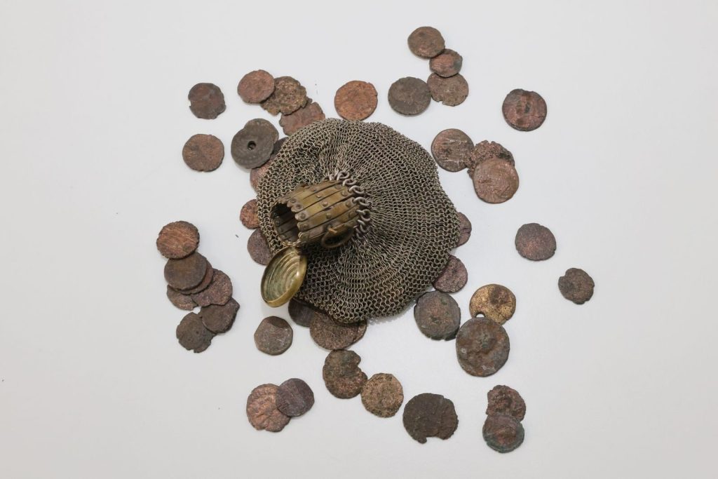 Top-down view of ancient coins scattered with a historical chainmail glove on a white surface.