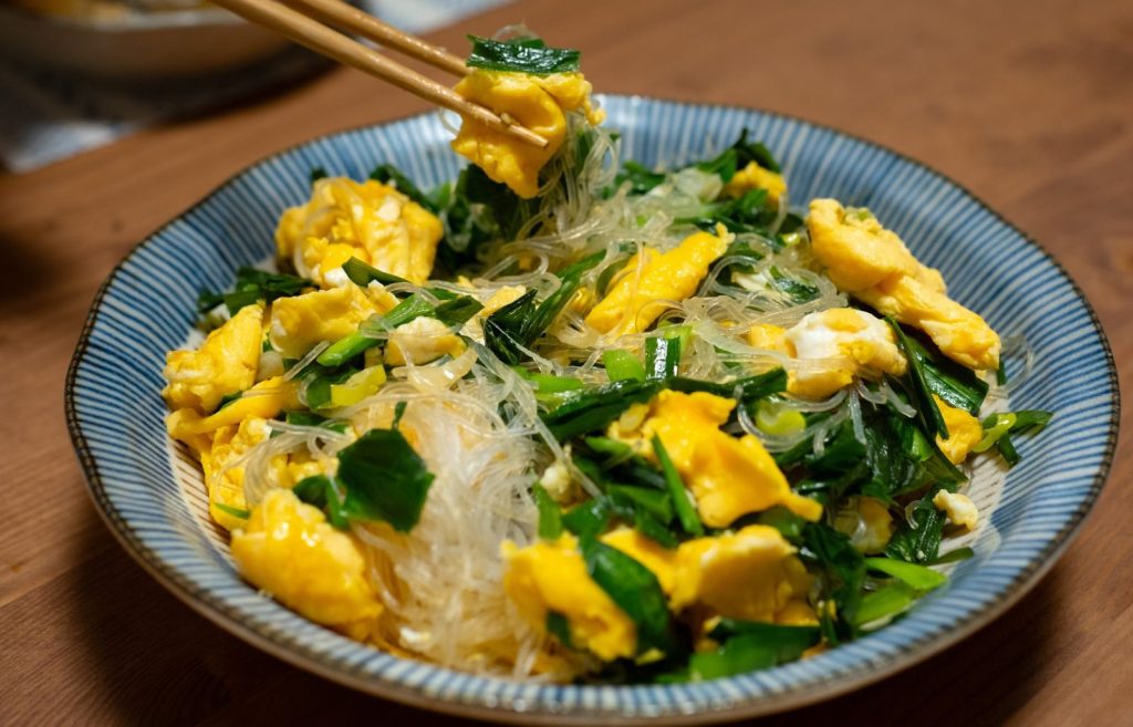 A vibrant Asian egg and vegetable stir-fry featuring noodles, captured indoors with a warm tone.
