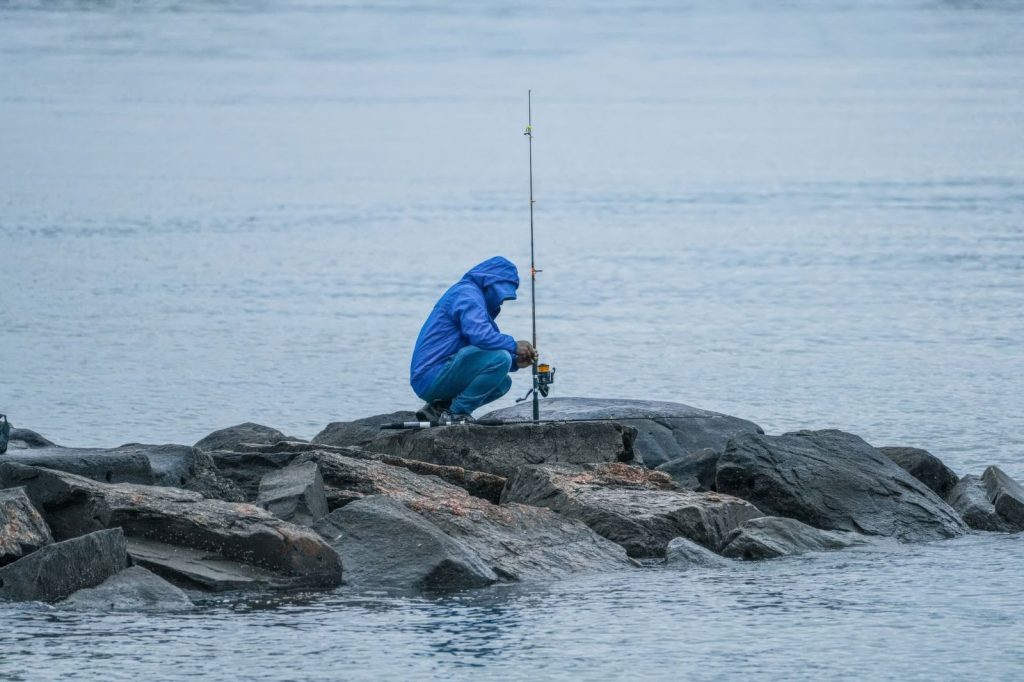 A lone fisherman in a blue jacket angles from rocky shore during overcast weather.
