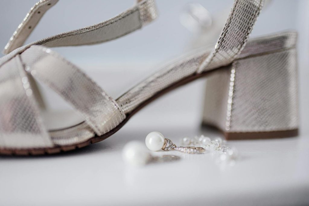 Close-up of bridal block heel sandals paired with pearl earrings on a white surface.