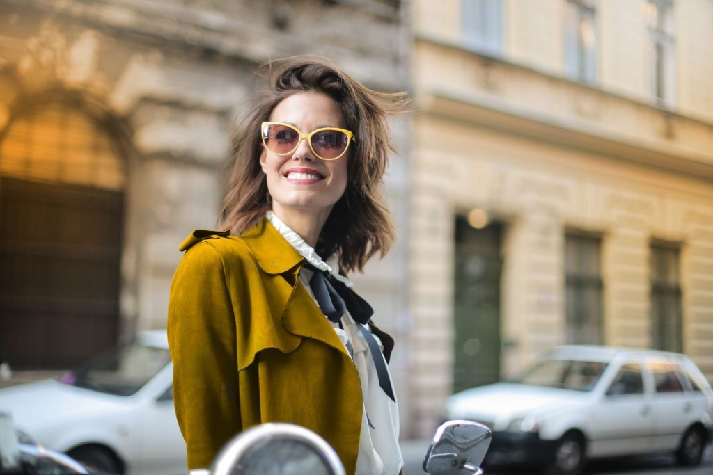 A woman in sunglasses smiles confidently in a chic yellow coat on a city street.