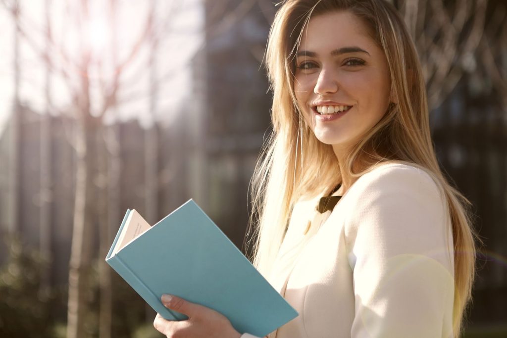 Cheerful young woman with blond hair holding a blue book and smiling outside during the day.