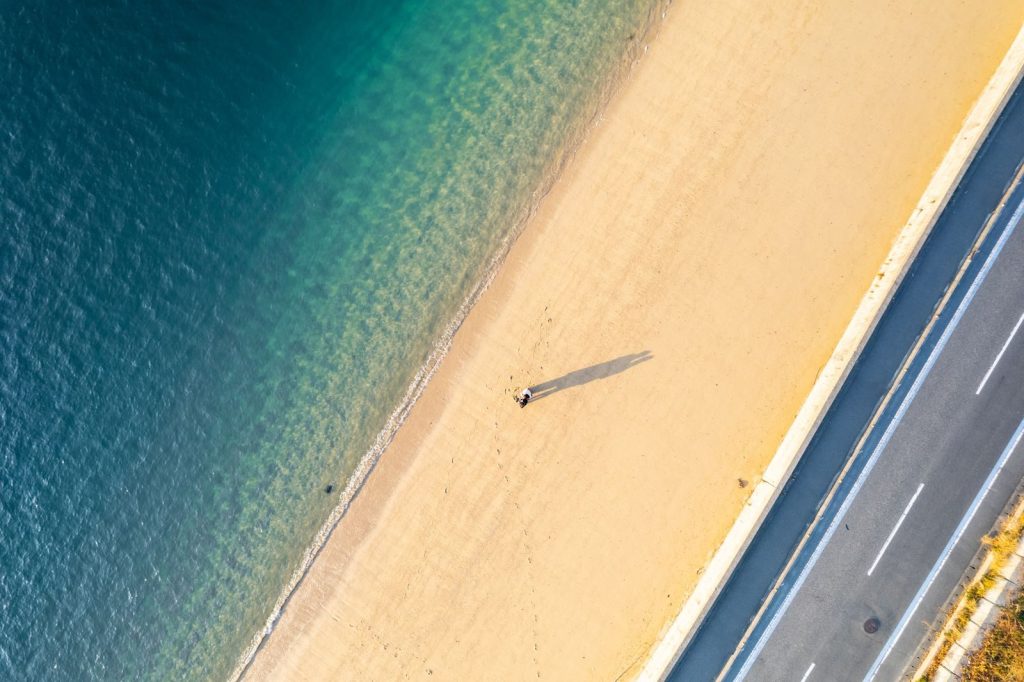 Aerial shot of a person walking alone on a sunny beach alongside a road, capturing serenity and solitude.