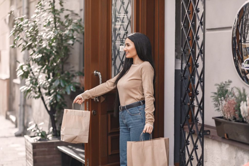 Side view of trendy young woman in stylish outfit carrying paper shopping bags while leaving boutique