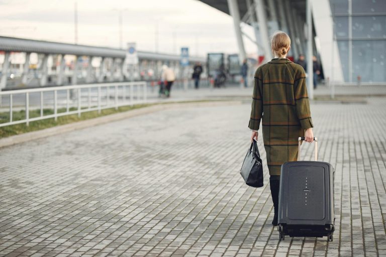 Back view full length of female executive in trendy outfit with luggage walking along sidewalk into airport building before departure