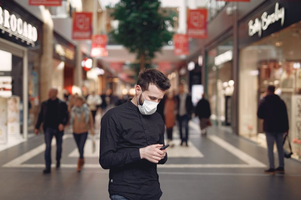 Bearded male student in casual clothes and mask messaging on mobile phone while strolling between stores in shopping mall